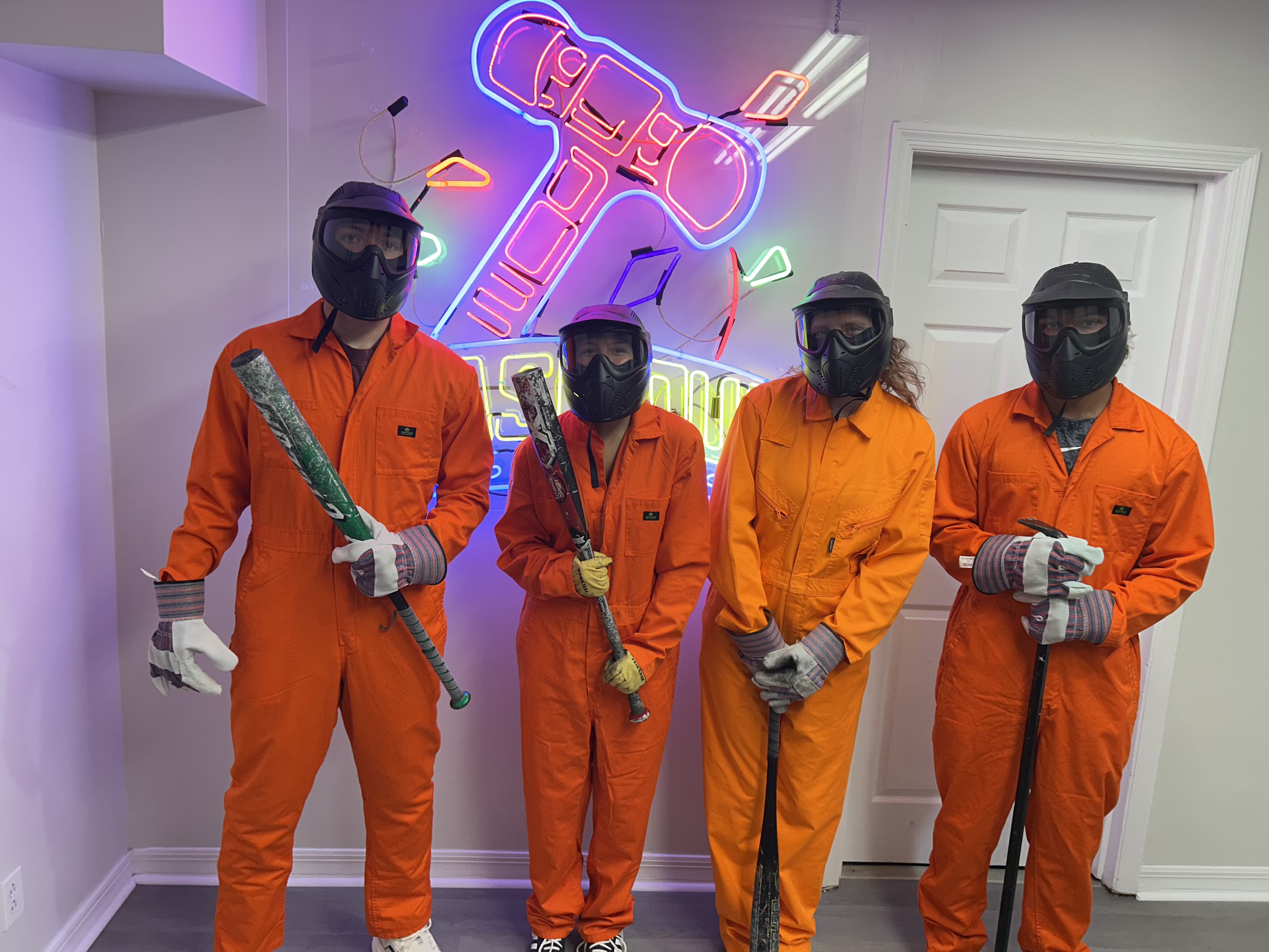 Crash Out customers in orange jumpsuits selecting baseball bats and smash tools from a barrel before their session