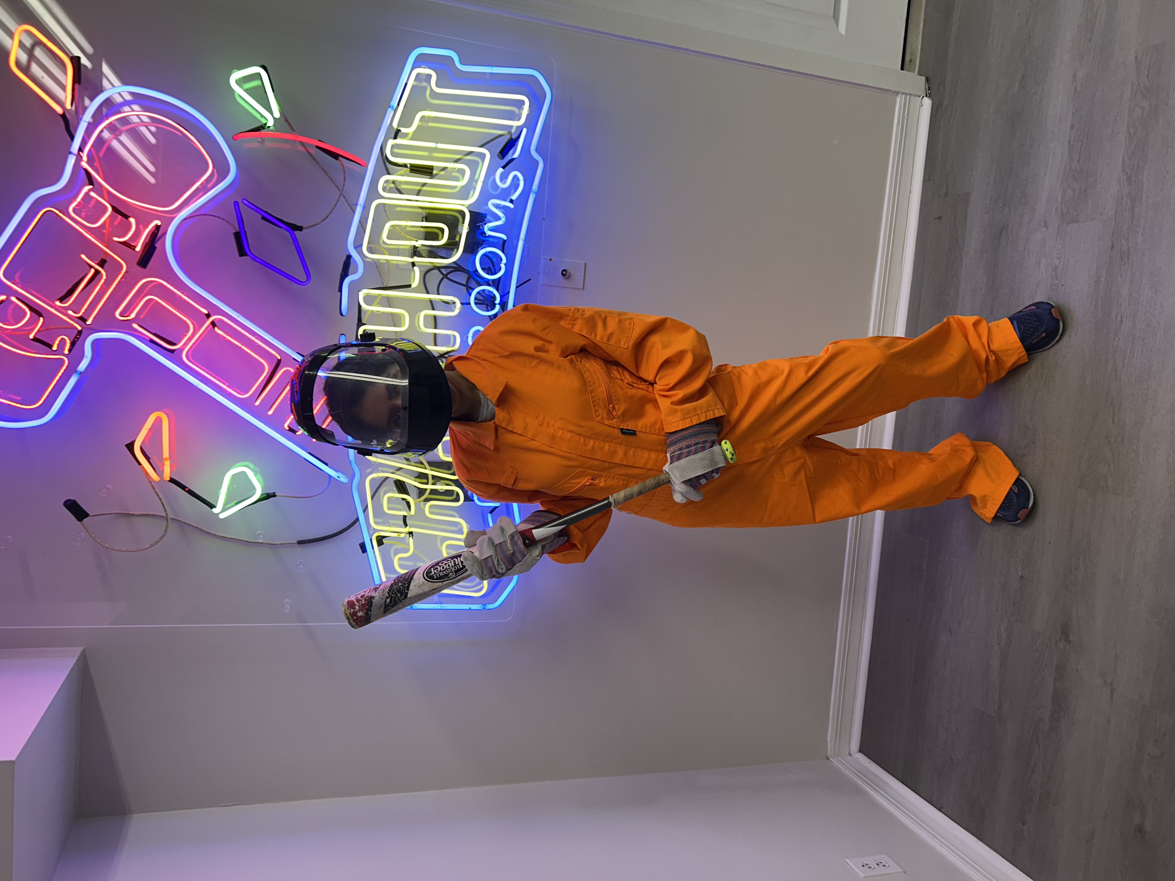 Solo Crash Out customer in orange jumpsuit and face mask posing with bat in front of colorful Crash Out neon sign