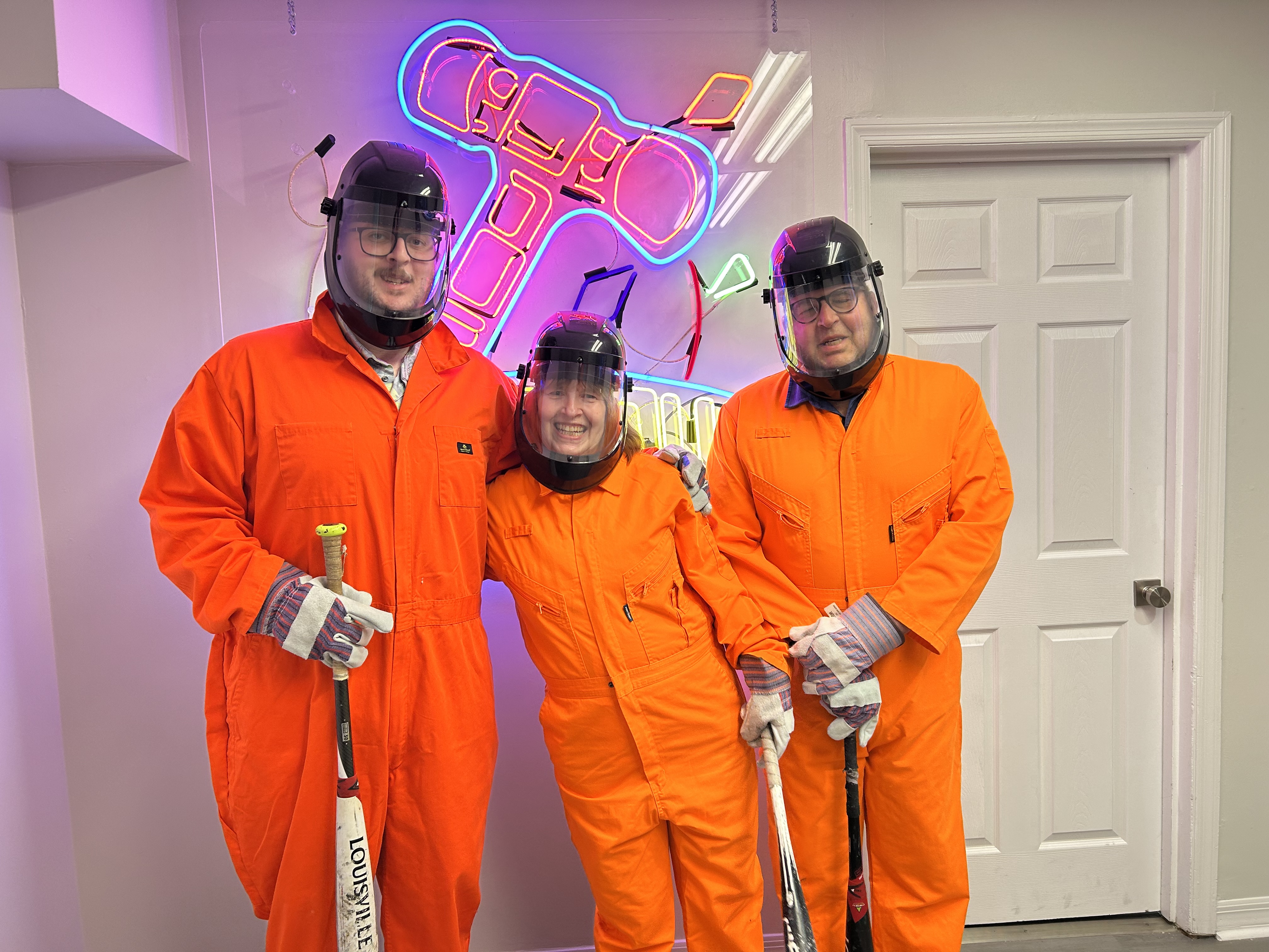Three smiling Crash Out guests in orange jumpsuits and face shields holding weapons before entering the smash room