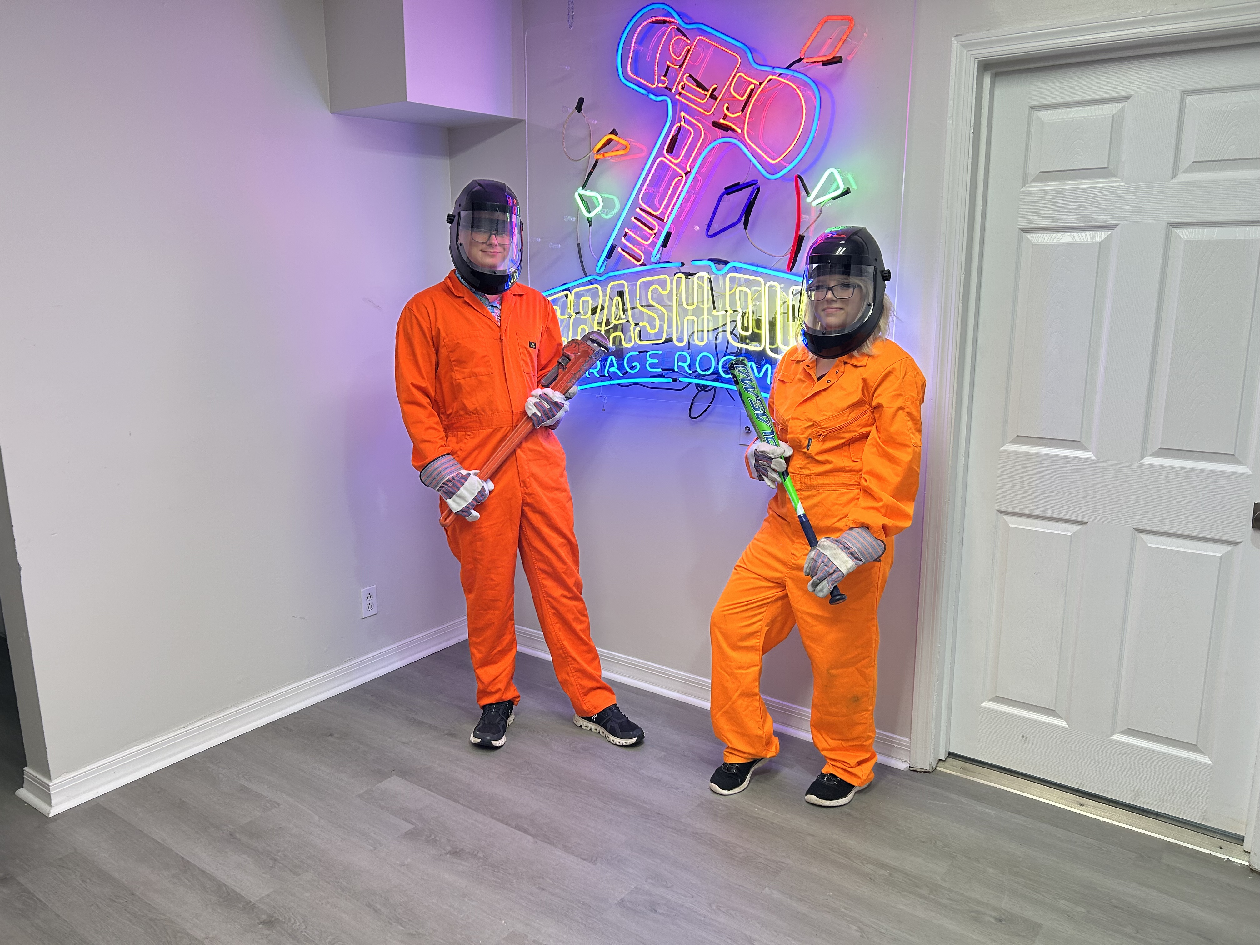 Large group of Crash Out Rage Rooms customers in orange jumpsuits holding bats and smash tools in front of neon sign