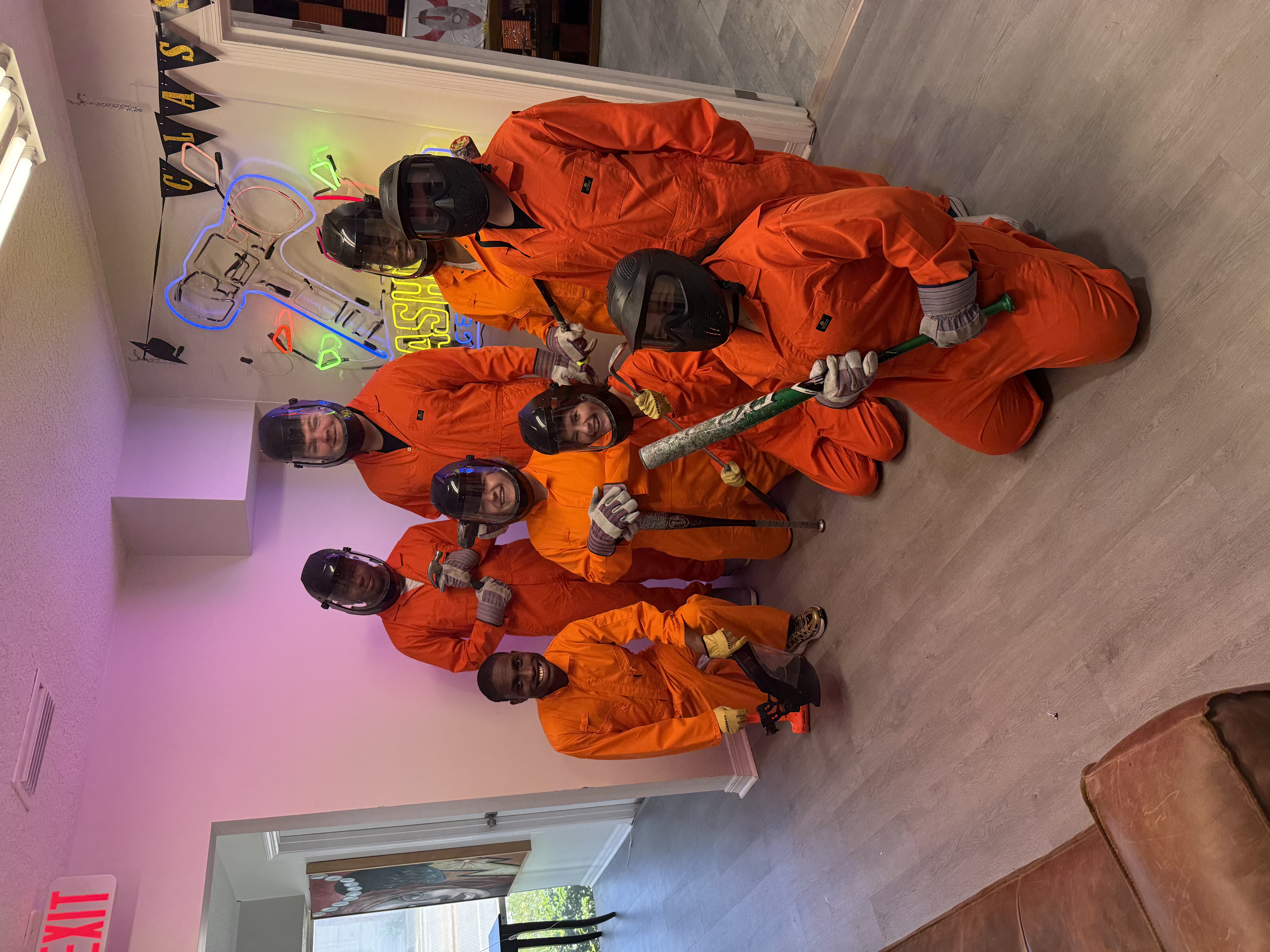 Eight Crash Out guests in orange protective jumpsuits posing together ready to enter the smash room